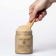 A hand holds a small bamboo scoop in an open glass jar of Little Seed Farm sugar scrub against white.