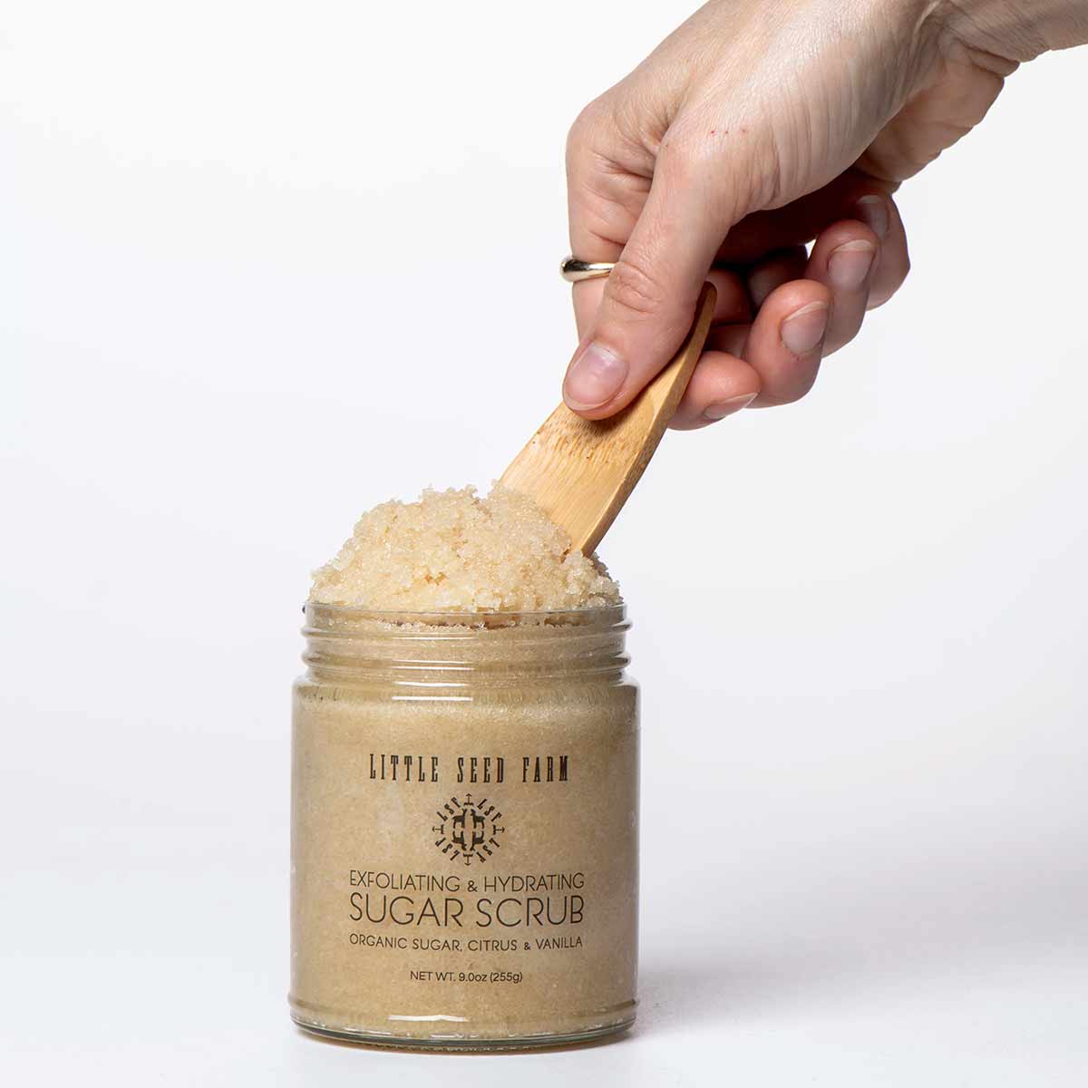 A hand holds a small bamboo scoop in an open glass jar of Little Seed Farm sugar scrub against white.