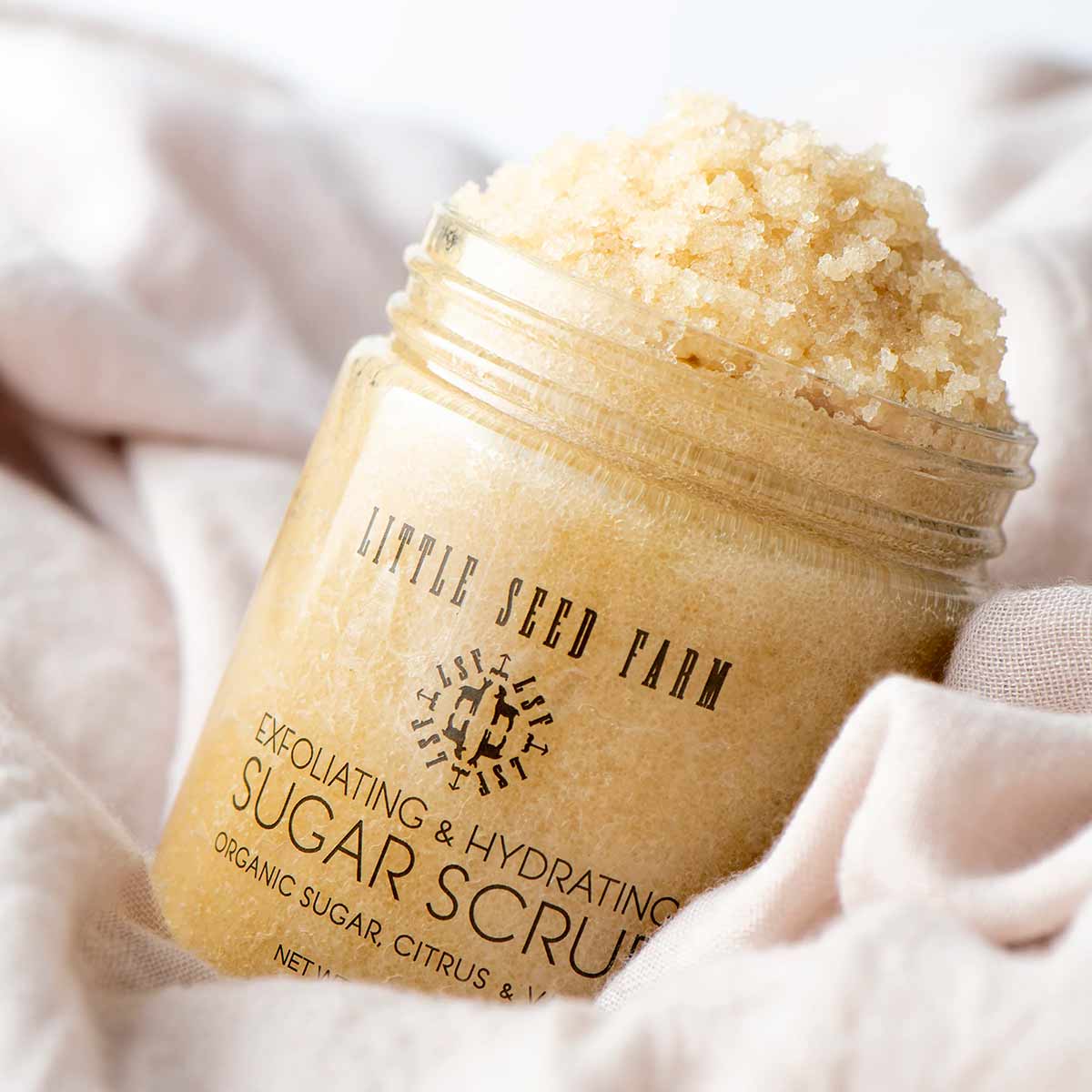 An open glass jar of golden sugar scrub with a Little Seed Farm label rests on soft cream fabric.