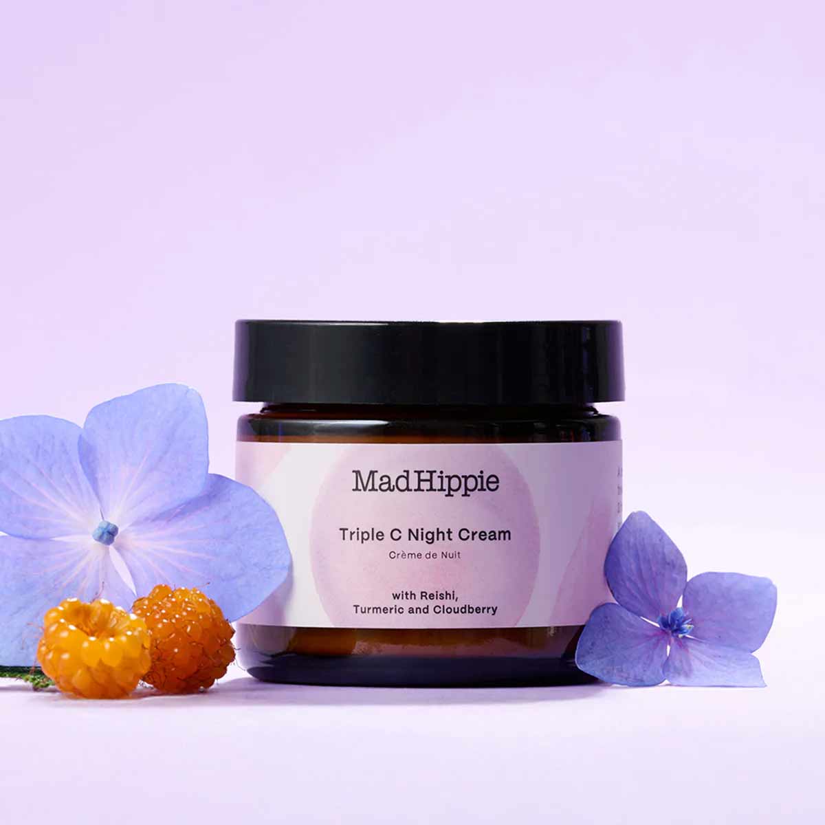 Mad Hippie Triple C Night Cream in amber glass jar with black lid, surrounded by blue flowers and orange berries on lavender 