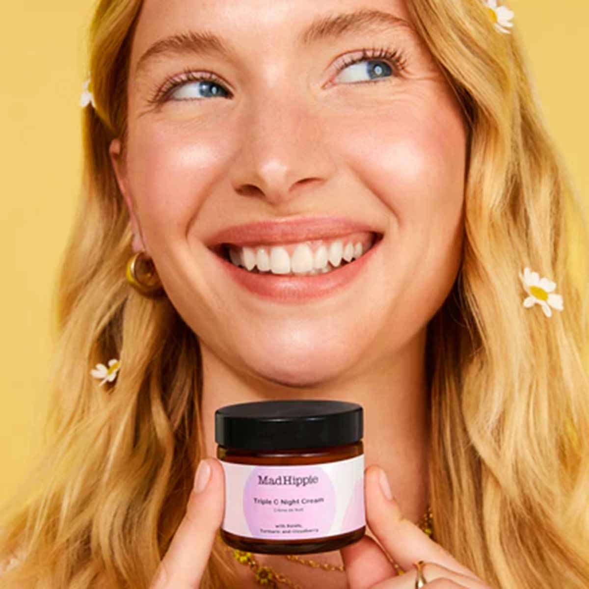 Woman with red hair holding Mad Hippie Triple C Night Cream jar with black lid against yellow background.