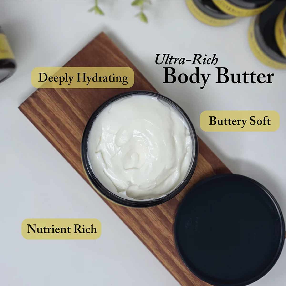 Open jar of white body butter with black lid on wooden board next to closed black jar.